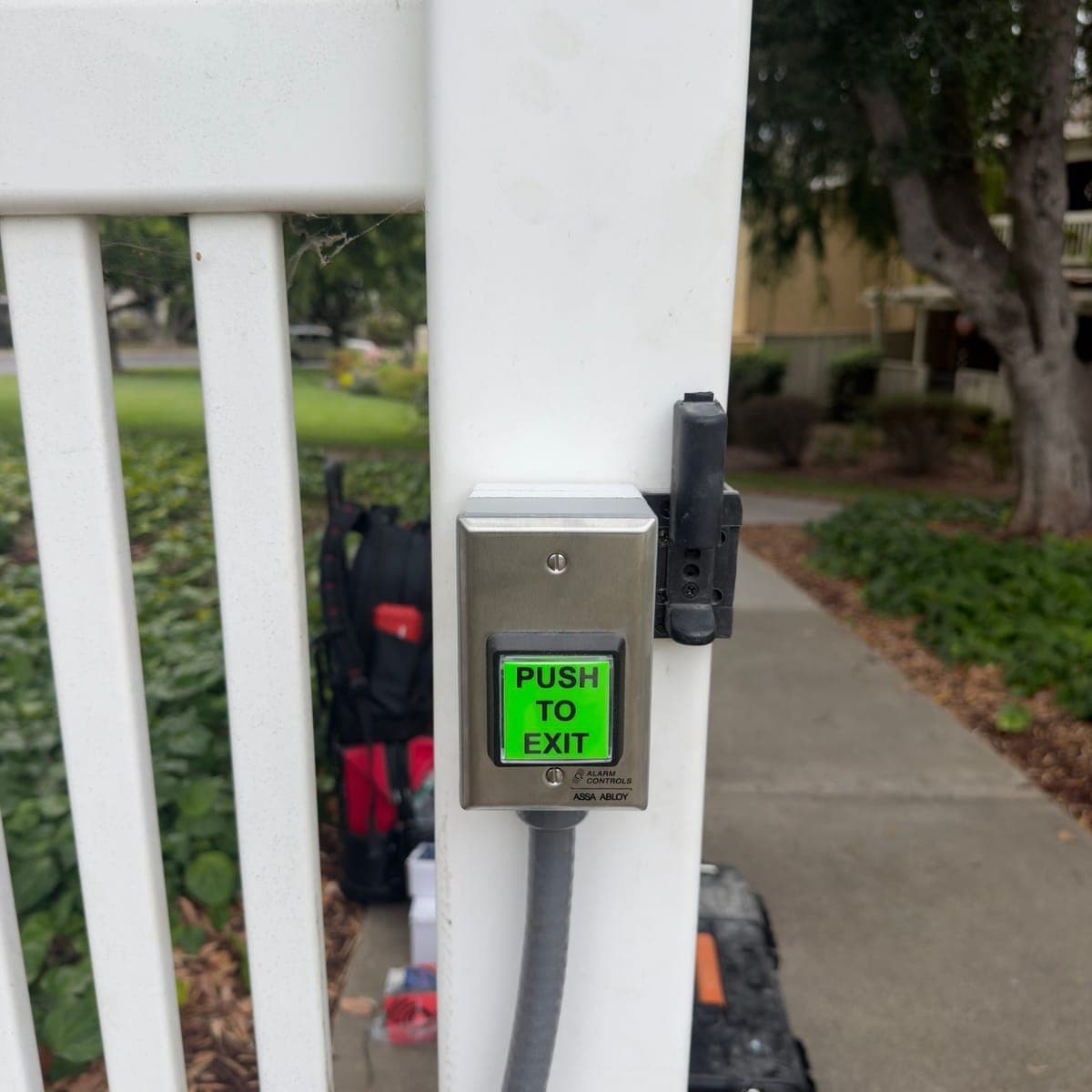 Green push-to-exit button in a stainless steel plate on a white gate post, with gray flexible conduit running down from it.