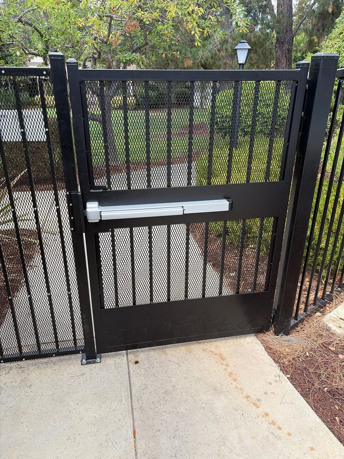Gray panic bar mounted across a black mesh amenity gate, with a concrete walkway, lawn and trees behind it.