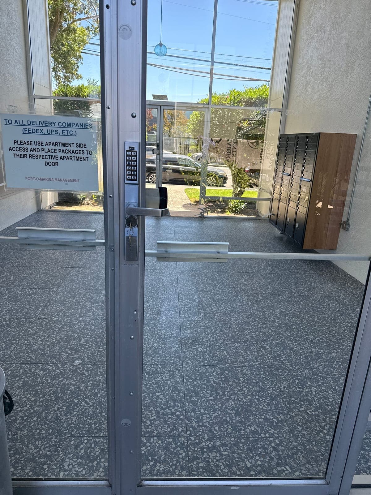 Aluminum and glass apartment entry door with a keypad lever trim on the stile, a delivery notice taped to the glass, and the lobby and mailboxes behind it.