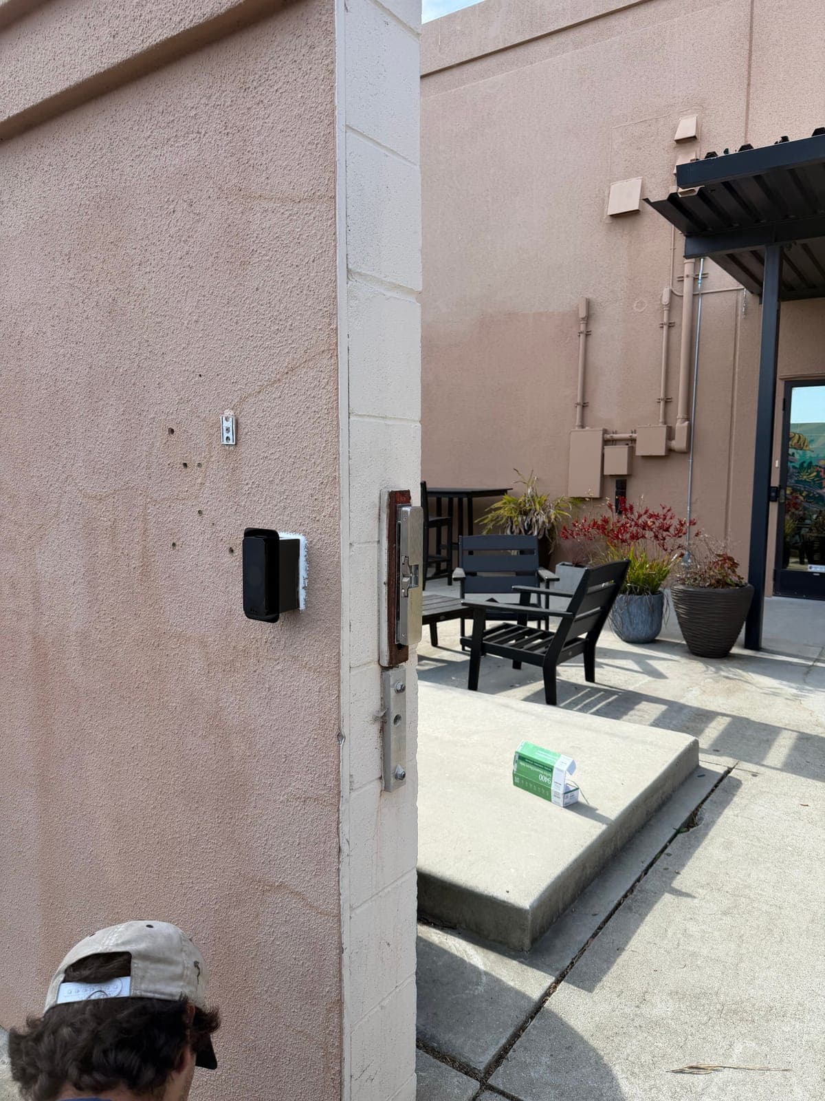 Card reader on a stucco wall beside a gate post fitted with electrified lock hardware, with a technician's cap at the bottom of the photo.
