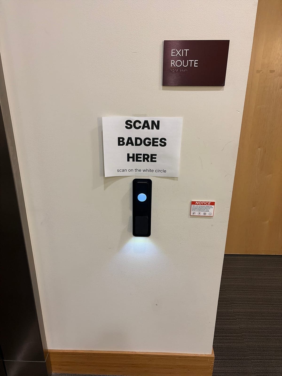 Black Kisi reader with a glowing white circle on a white wall next to a brushed stainless steel panel, under a paper sign reading 'Scan badges here' and a maroon 'Exit route' sign.