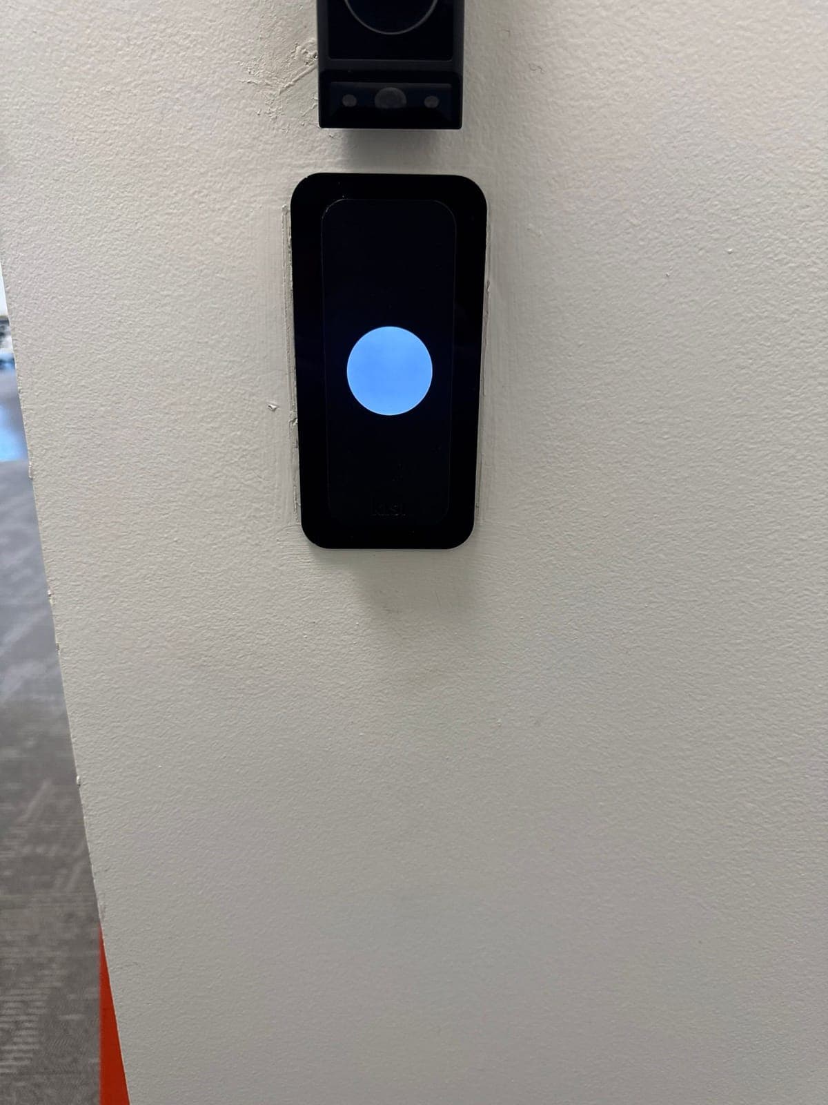 A Kisi card reader mounted on a white office entry wall, with the bottom edge of a video doorbell visible above it.