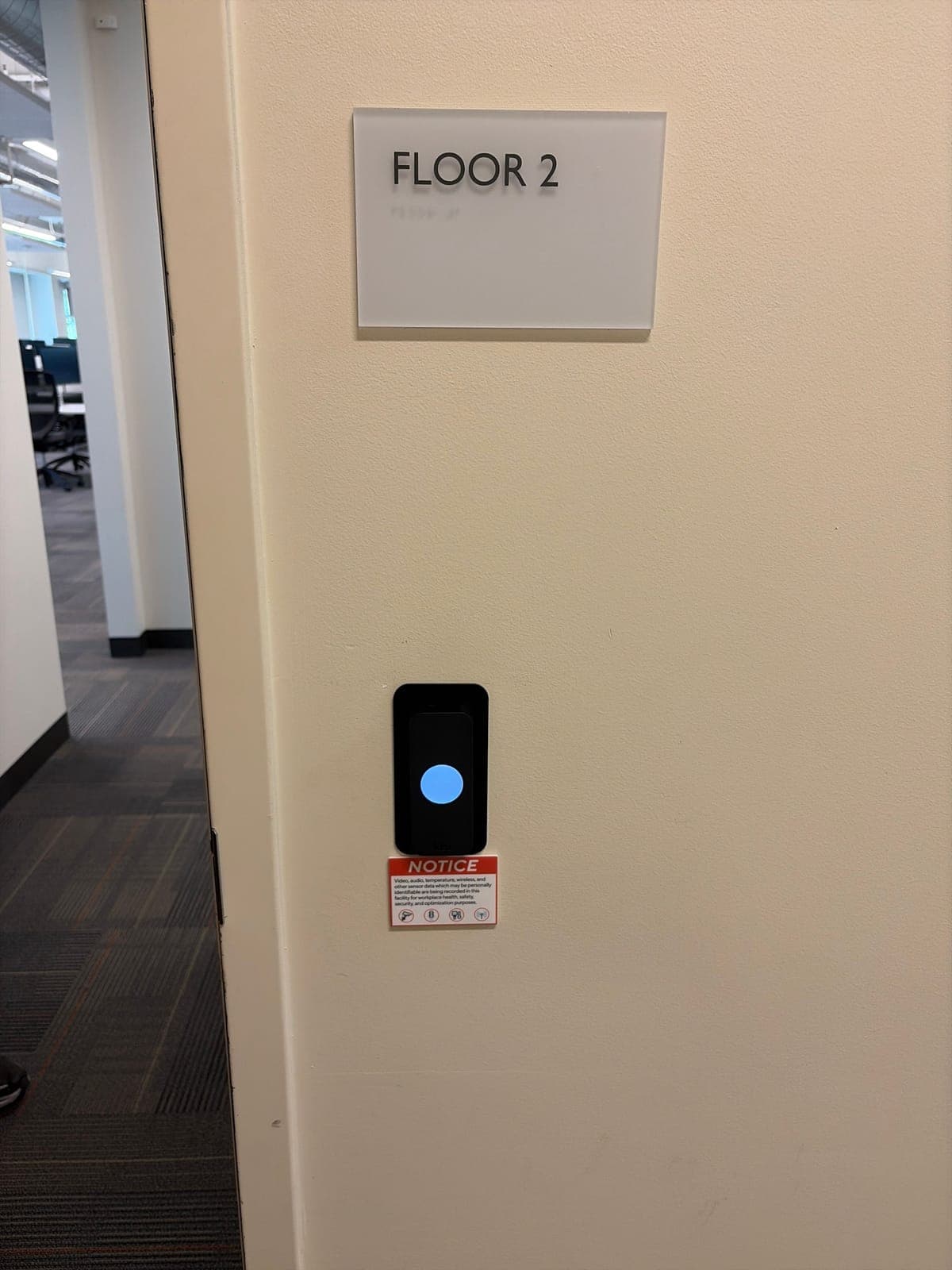 Close-up of a black Kisi card reader on a cream wall under a Floor 2 sign, with an open-plan office visible through the doorway.