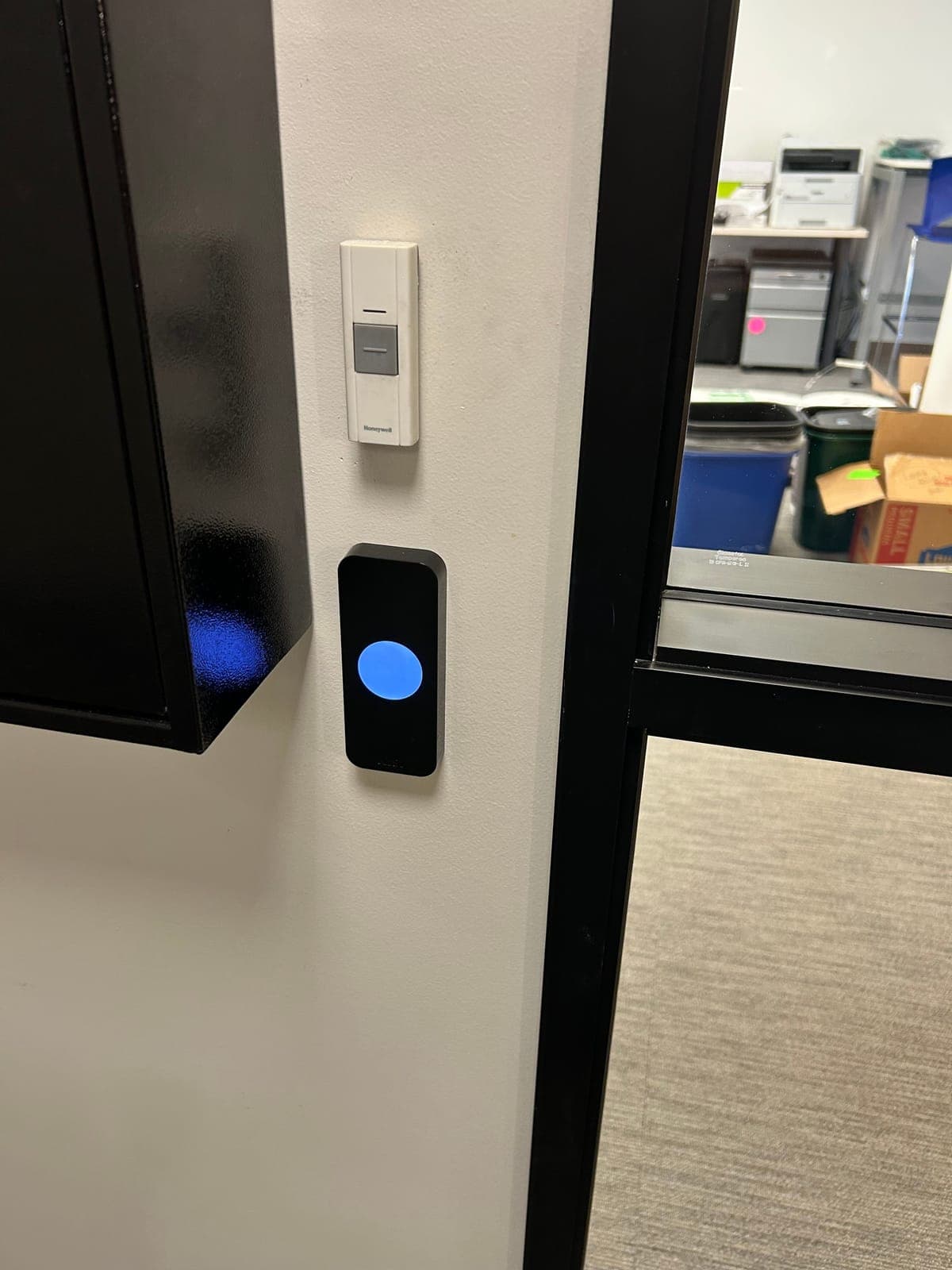 Black Kisi reader with a lit blue circle mounted on a white wall beside a dark-framed glass office door, with a doorbell button above it.