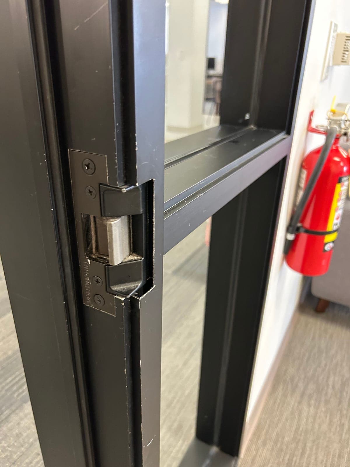 Von Duprin electric strike installed in a black aluminum office door frame, with a glass sidelite and a fire extinguisher in the background.