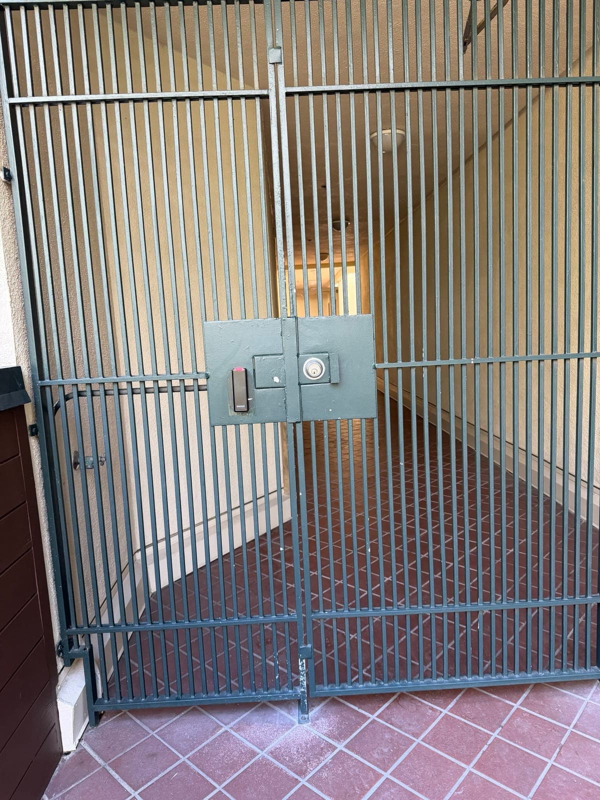 A green steel bar gate in a tiled passage, with a small black reader and a round key cylinder mounted on square lock plates at mid-height.