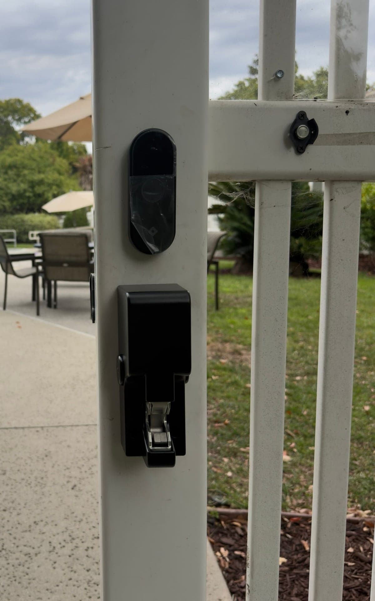 A black oval reader with protective film still on it, mounted above a black surface-mount gate lock on a white metal pool gate post, with patio chairs and umbrellas in the background.