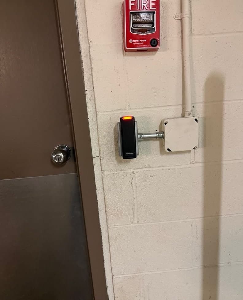 Before and after: a black rounded reader on a conduit stub from a surface junction box on a painted block wall, then a smaller black ButterflyMX reader on the same conduit stub and box beside a brown door, with a red fire alarm pull station above.