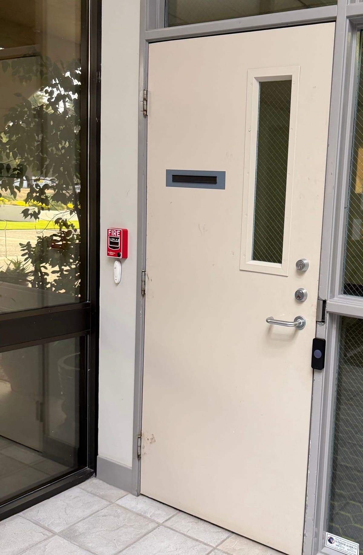 A card reader on the aluminum frame beside a steel staff door with a narrow vision panel, next to a fire alarm pull station.