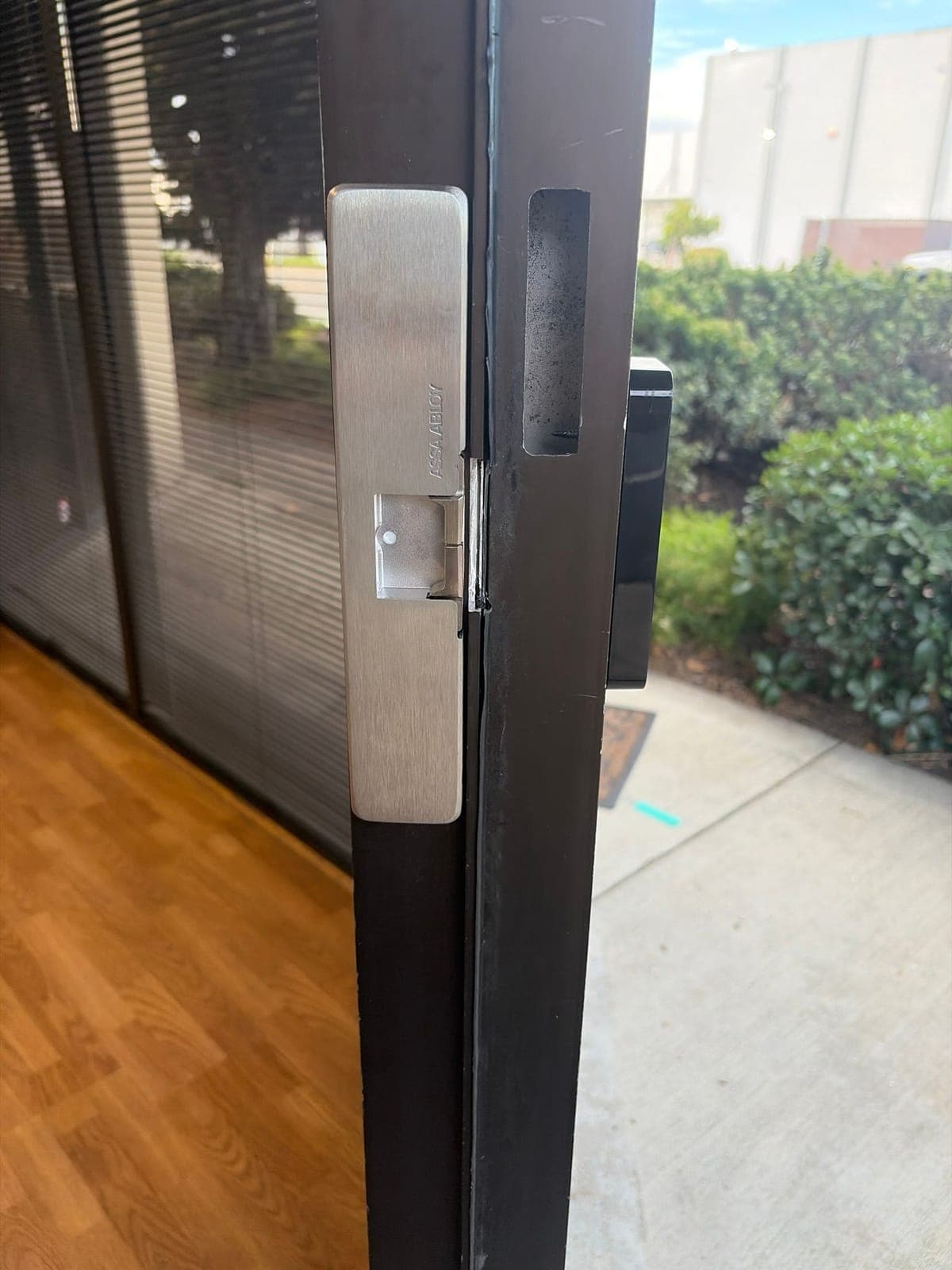 Brushed steel ASSA ABLOY electric strike installed in the edge of a dark aluminum storefront door frame.