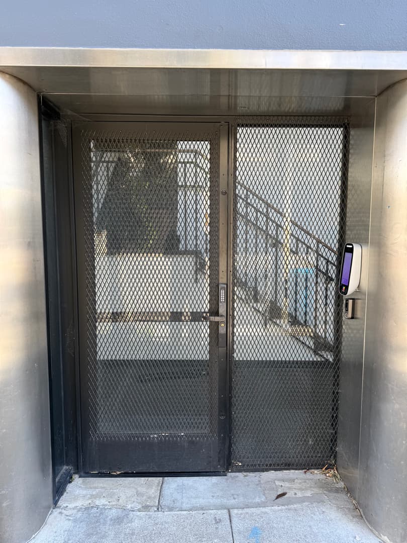 Controlled building entry with a mounted video intercom beside a metal mesh door.