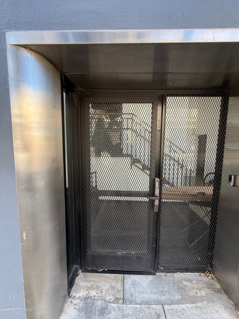 Exterior metal mesh entry door with keypad access and an intercom mounted near the frame.